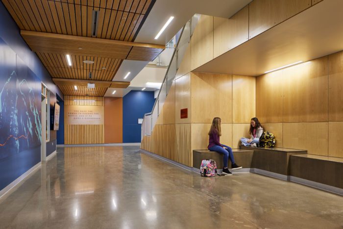 Front hallway of Yelm Middle School shows students in a sitting area to the right, under the stairs. On the left, wood features are seen along the ceilings and walls along with deep blue wall finishes.
