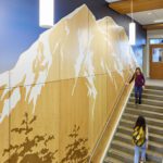 students climb the large front staircase of Yelm Middle School. To the left of the staircase, a two-story graphic of Mt. Rainier in a wood finish and pine trees in a brown relief with a dark blue sky create a bold feature. Wood slats are also featured in periodically along the ceiling, and long white pendant lights illuminate the stairs.