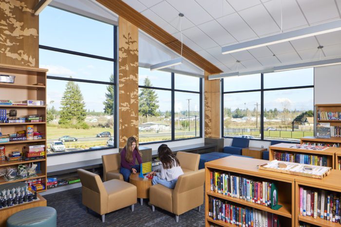 the library of Yelm Middle School boasts wide windows with views to Mt. Rainier and graphics of pine trees. Students are seated in cushioned chairs, facing each other. Low bookshelves are seen in the foreground.