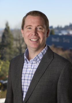 Headshot of Joseph Rydman in a suit jacket and plaid button-down shirt smiling at the camera.