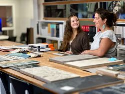 BCRA Intern Gabriella Pearson and Interior Designer at BCRA, Marissa Rosati, converse at a design table with a variety of design samples on the surface of the table Gabriella is smiling.