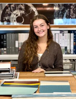 BCRA Intern Gabriella Pearson smiles at the camera in front of a work table at BCRA's office